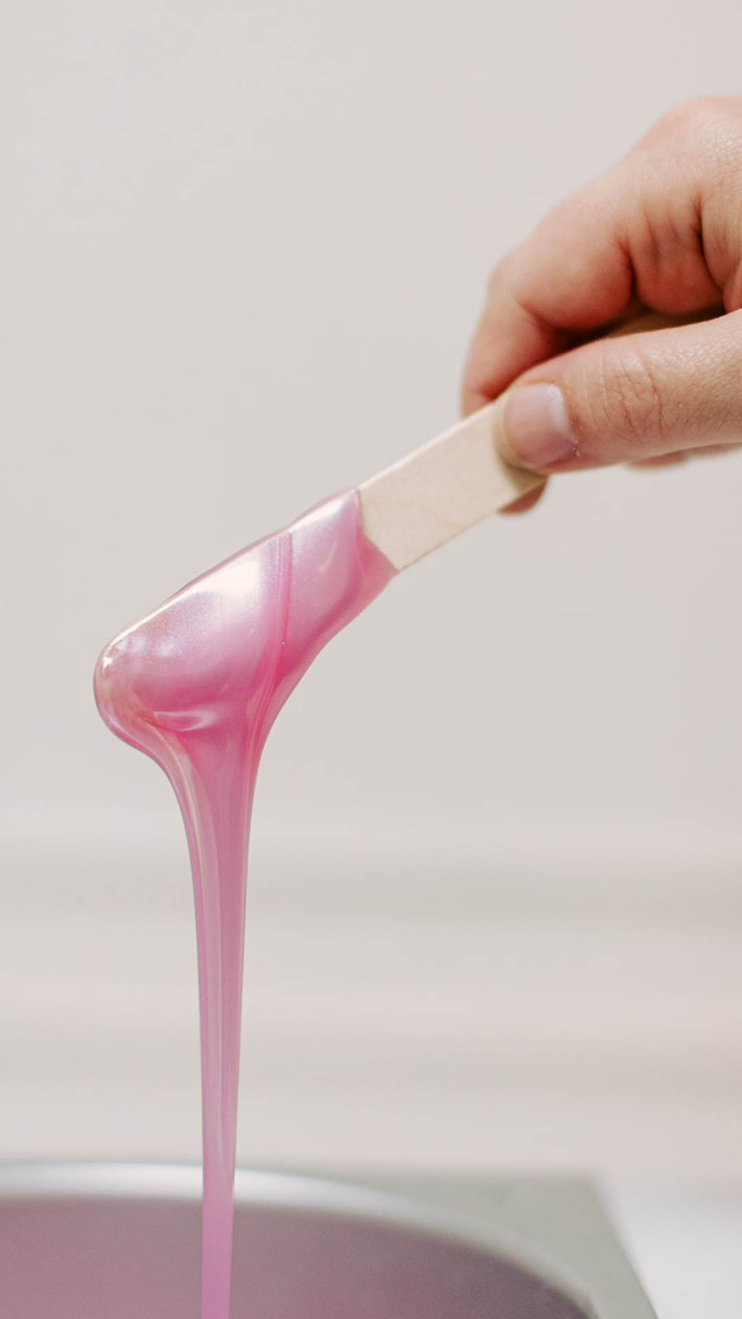 Hand holding a waxing stick over a wax pot with pink wax dripping.